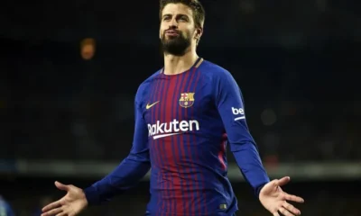 Gerard Piqué standing on the pitch in a Barcelona jersey, raising his hand with a serious expression.