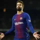 Gerard Piqué standing on the pitch in a Barcelona jersey, raising his hand with a serious expression.