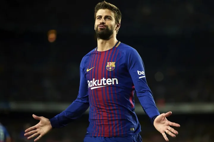 Gerard Piqué standing on the pitch in a Barcelona jersey, raising his hand with a serious expression.